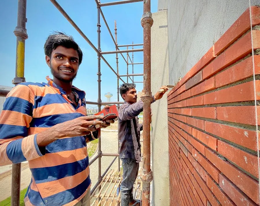 A crew leader on the phone beside a mason laying bricks on scaffolding